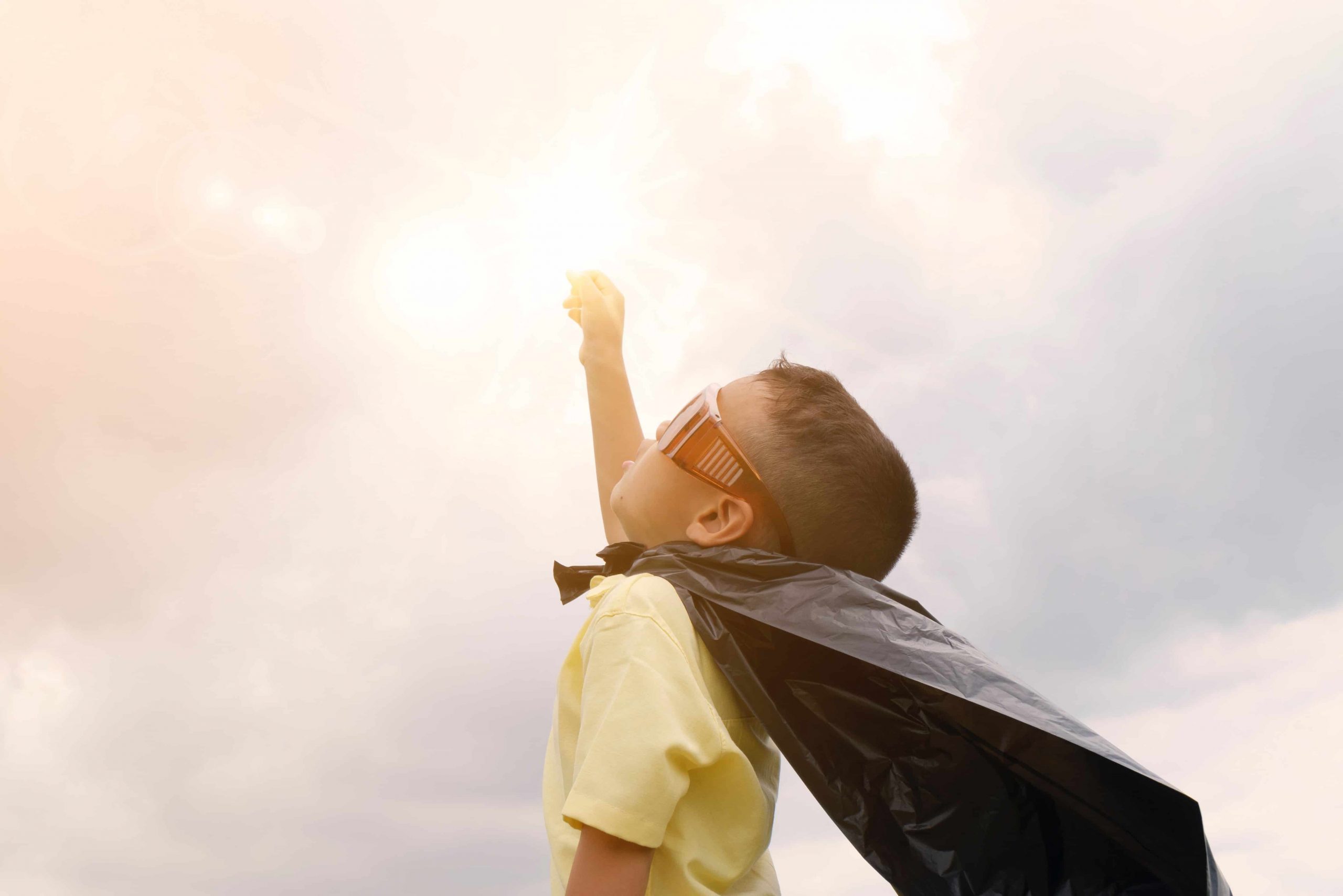 A boy dressed in glasses and a garbage bag cape pointing his fist towards the sunny sky