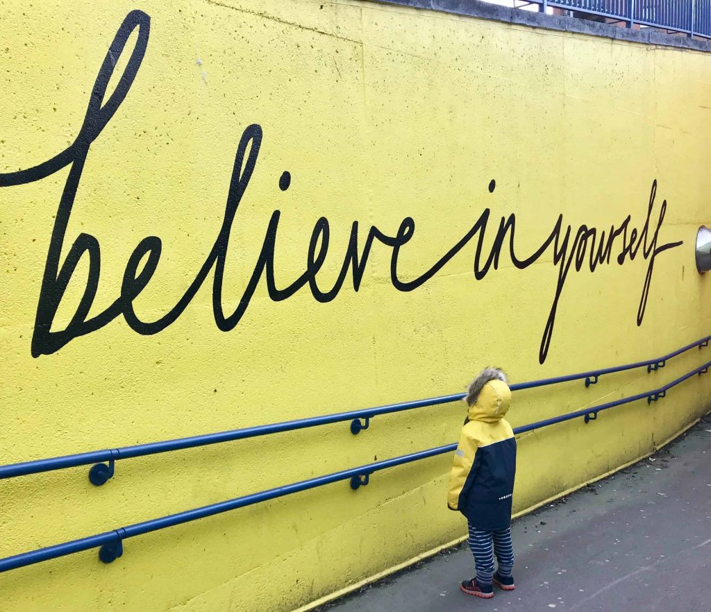 Child looking at a yellow wall with "believe in yourself" painted on it