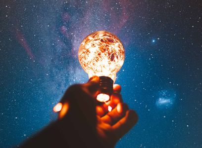 A hand holding a light bulb filled with string lights against the starry sky