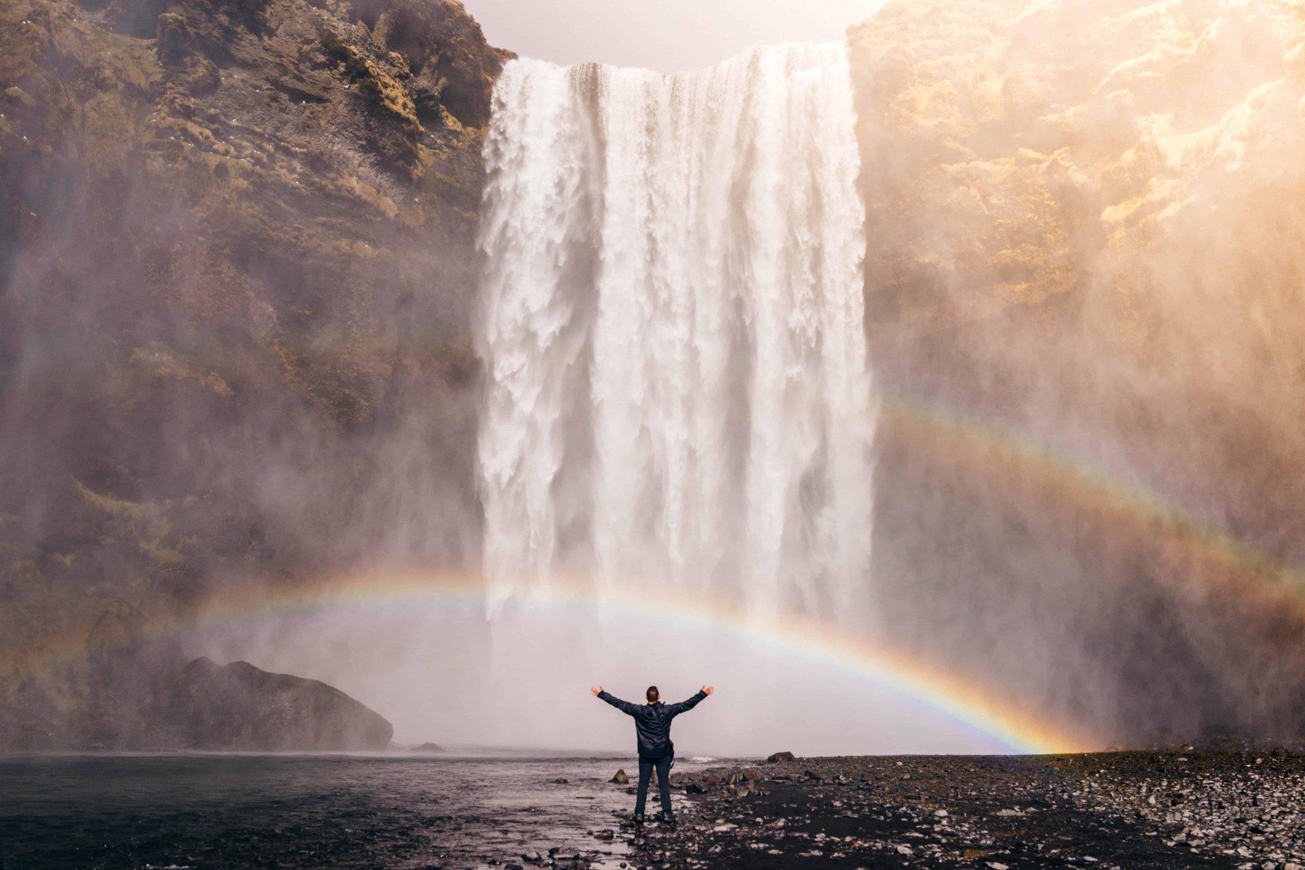 Person standing at the base of a waterfall over large rocks and a double rainbow
