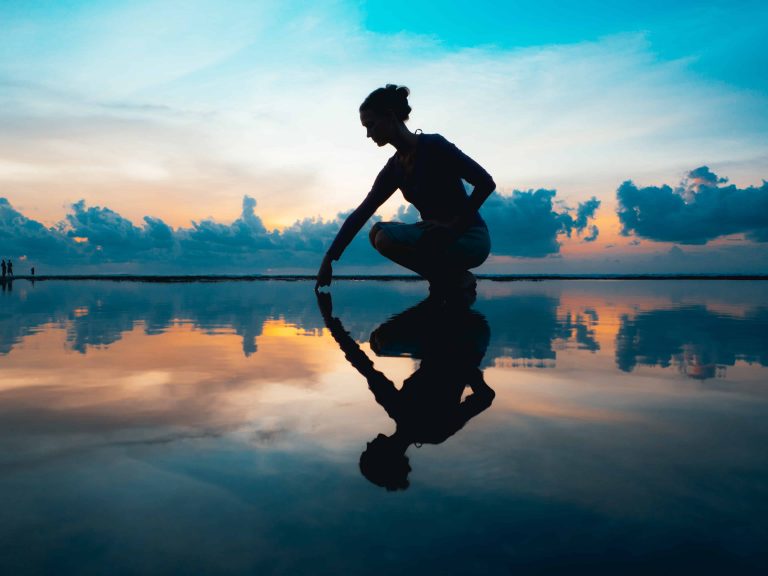 A woman bending town to touch her reflection in the water with low clouds and sun behind her