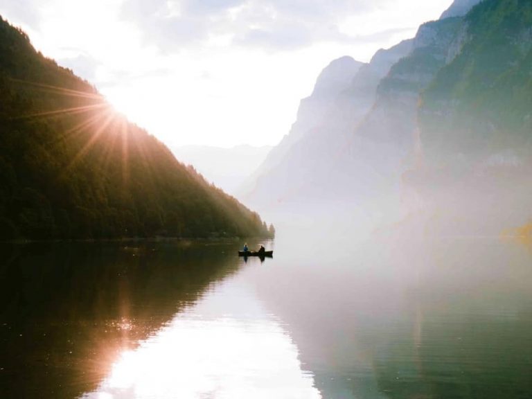 Two people in a row boat on a calm lake surrounded by mountains and a sunny sky
