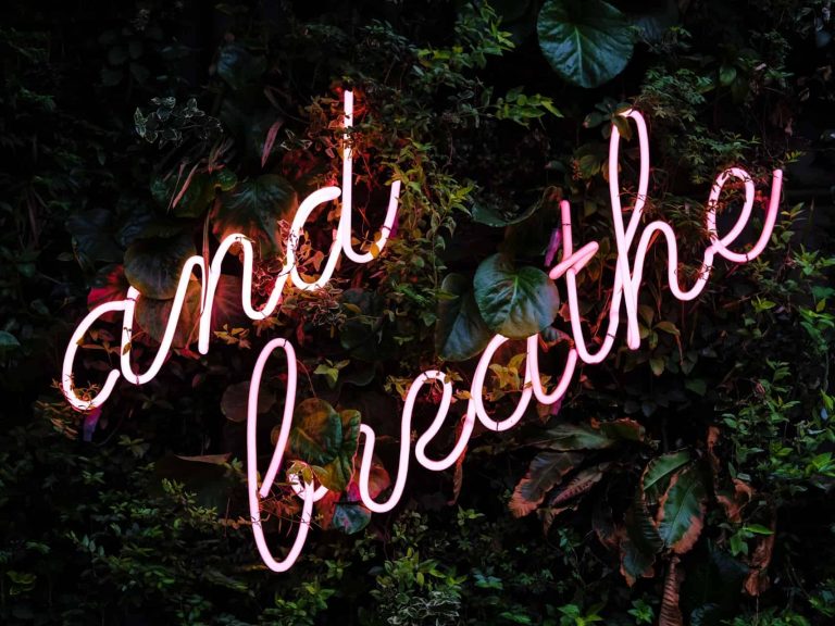 "And breathe" sign in neon lights against wall of leaves