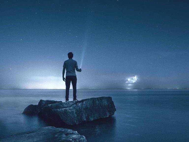 A person standing on a rock in the water with a flashlight at night