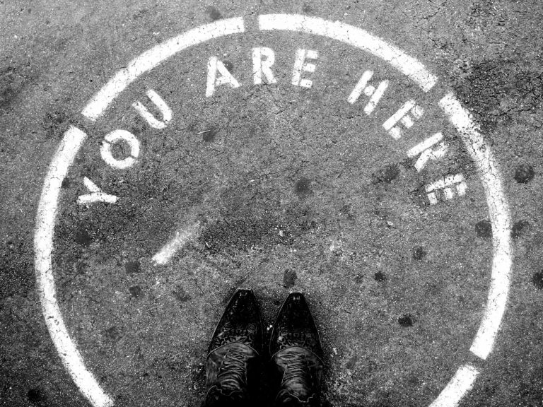 A person standing on the sidewalk with their feet in a painted circle that says "You are here"